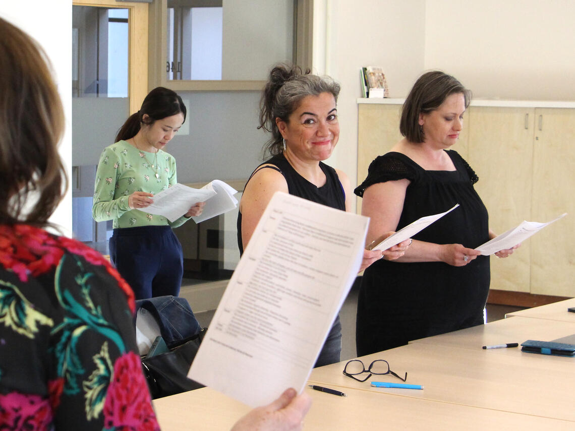 A group of women stand around a table reading from scripts