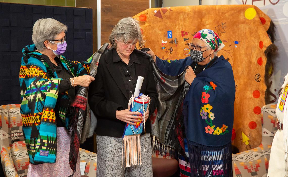 Former Provost Dr. Dru Marshall and Elder Rose Crowshoe drape Provost Teri Balser with a shawl to commemorate the transition of the strategy during the transfer ceremony to celebrate the fourth anniversary of  ii’ taa’poh’to’p.