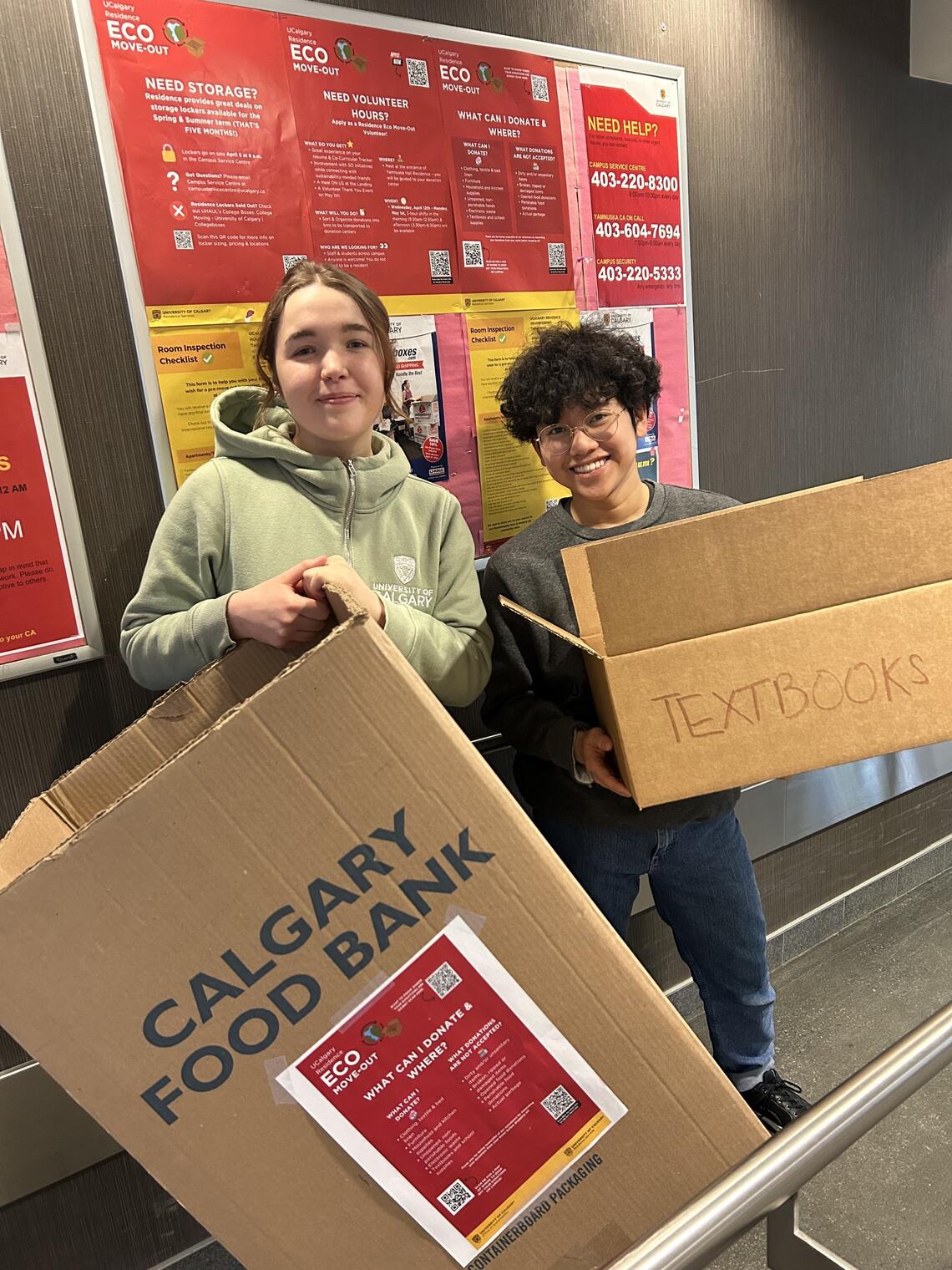 Two people stand smiling at the camera holding boxes labelled 'Calgary Food Bank' 