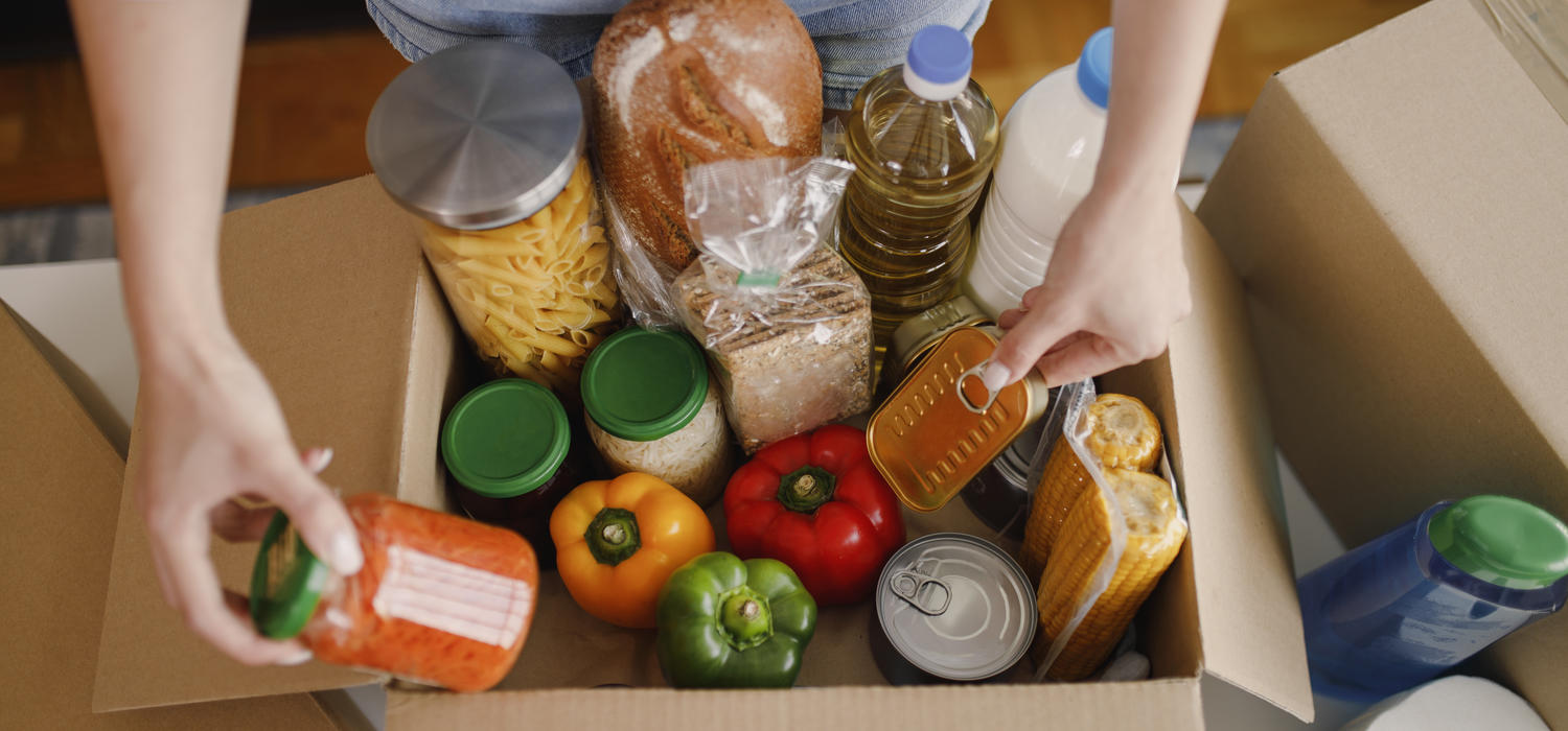 Person standing at a table with a box of food, interacting with a jar and a can of food inside the box.