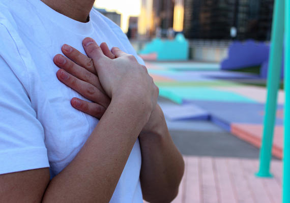 A man in a white t-shirt standing in a playground with arms folded over his chest; his face is outside of the frame