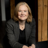 Portrait style photo of Mary Brindle wearing a suit jacket and leaning on a chair
