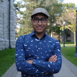 a brown man, wearing a hat and round black glasses, wearing a blue shirt and smiling toward the camera