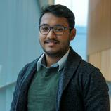 Viki Kumar Prasad wearing glasses, a green sweater, and a gray jacket, standing indoors with a softly lit background of windows and wooden paneling