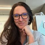 Close up image of Dr. Shaw's smiling face. They have long brown hair and are wearing navy glasses and a light grey blazer. They are resting their chin on their hand.