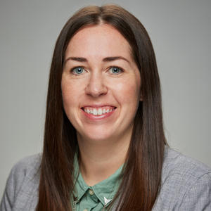 White woman with brown hair, smiling at the camera, wearing a green shirt and grey blazer against a grey background.