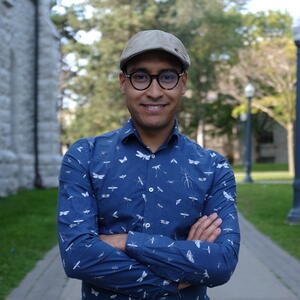 a brown man, wearing a hat and round black glasses, wearing a blue shirt and smiling toward the camera