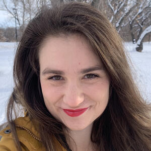 Headshot of a woman with long brown hair and brown eyes in a yellow parka against a snowy backdrop.