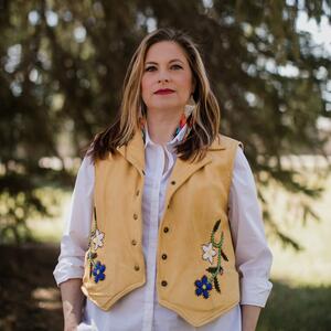 Dr. Melanie Griffith Brice is wearing a beaded Métis vest. She is standing in front of trees. 